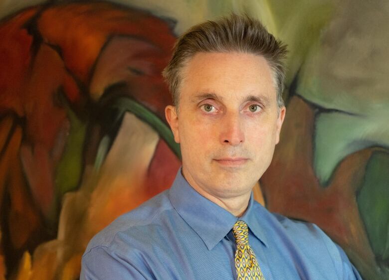 A man wearing a dress shirt and tie poses for the camera. A colourful painting is in the background.