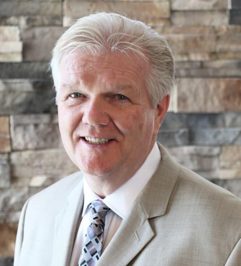 A man with white hair wearing a grey suit and purple tie smiles in front of a brick wall.