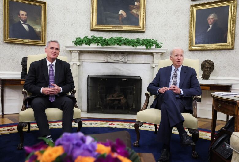 Two men in suits sit in arm chairs with fireplace between them and portriats of past US presidents on the wall