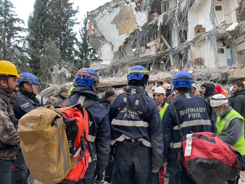 Firefighters and others working to rescue people trapped in a collapsed building stand in front of rubble.