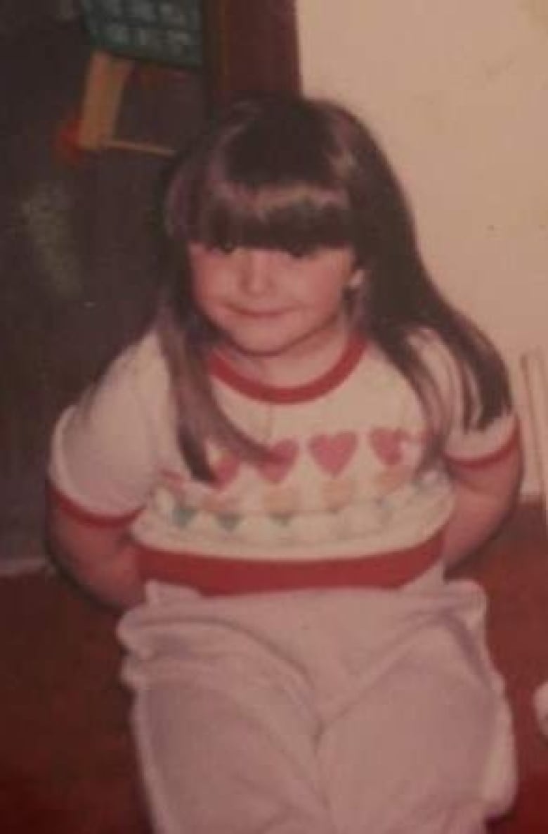 An old colour photograph shows a young girl, wearing a t-shirt with hearts on it and pajama pants. She is smiling and holding her hands behind her back.