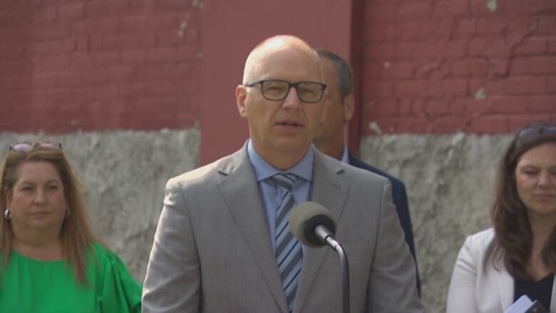 A bald man in a grey suit stands at a microphone as people stand behind him and to his sides.