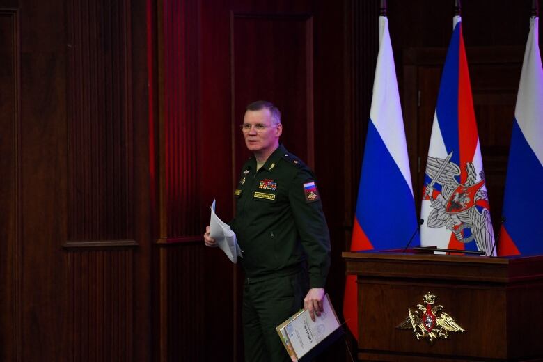 A person in military officer garb holding papers walks away from a lectern, behind which are seen Russian flags.