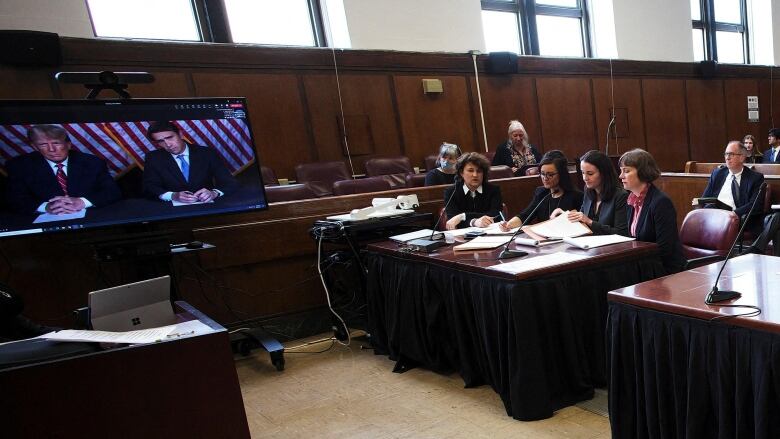 Donald Trump is seen on a video screen seated beside another man, in a court room