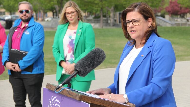 A woman in a blue blazer speaks behind the microphone at a news conference, while two other people stand behind them.