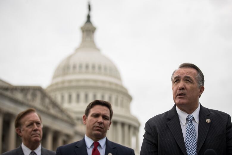 Three men in suits are shown speaking outside in front of the U.S. Capitol.