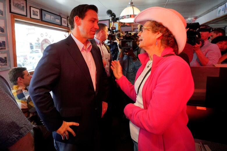 A man in a blazer and collared shirt listens to older woman in a cowboy hat speakng to him.