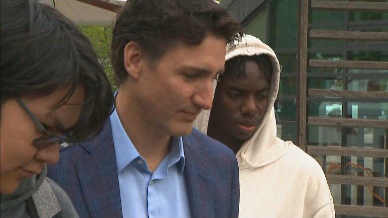 A man in dark hair and a suit looks off to the right of the photo frame. A male teen with black hair and glasses stands at the left of the photo while another student, wearing a white hooded sweatshirt stands at the right.