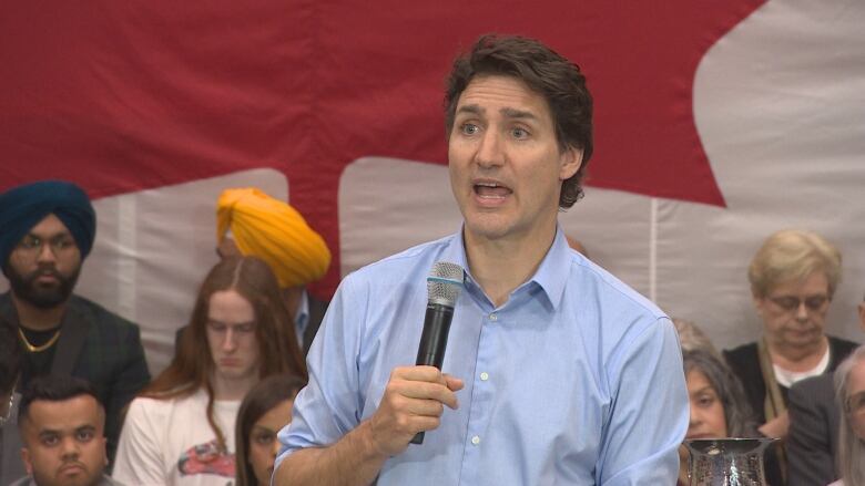 A man with dark hair sits at a table covered in a dark tablecloth in front of a row of people sitting on risers. Behing them all is a Canada flag.