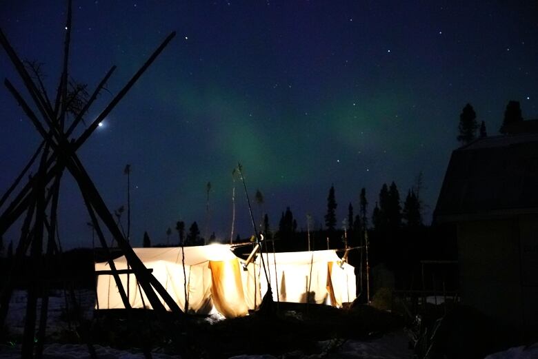 The light from inside two white canvas tents glows under a dark night sky with the aurora borealis faintly gleaming through.