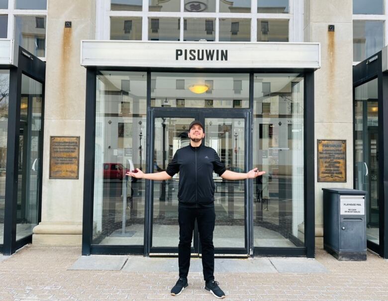 A man stands in front of a theatre building, looking up at the sky with his arms spread wide.