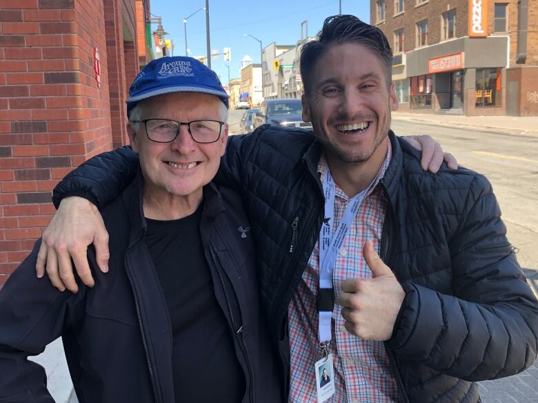 Two men smiling while standing outside next to a downtown street.