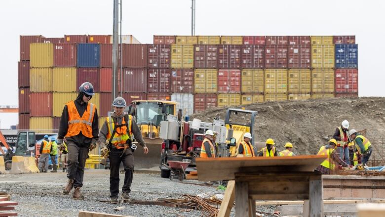 Two men in reflective vests and hard hats walk through a construction site with containers in the background