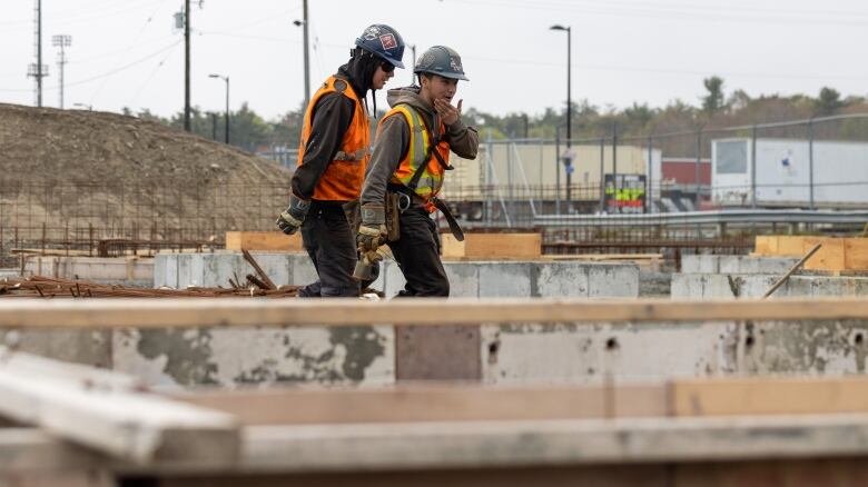 Two workers in orange vests and hardhats walk by piles of lumber