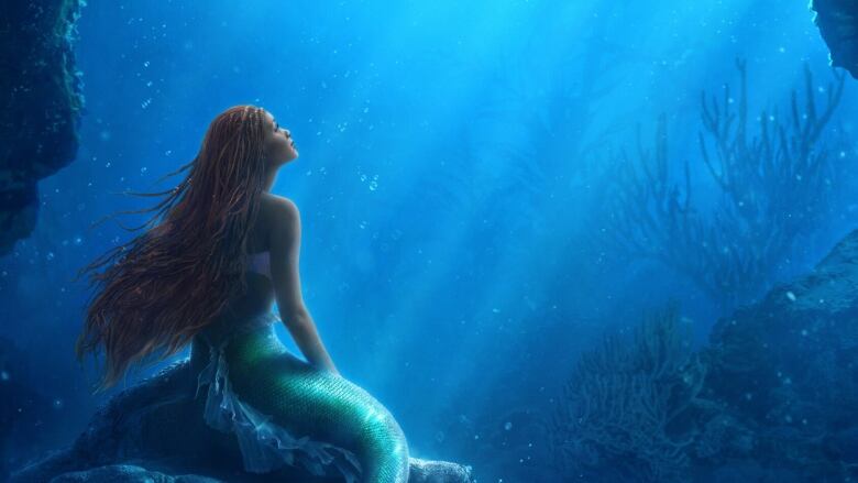 A mermaid woman sits on a rock under the ocean. She is looking up to the light above her. 