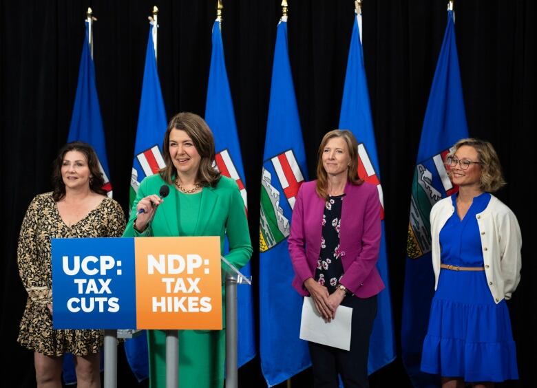 A woman speaks at a podium, flanked by three other women. A series of Alberta flags is behind them.