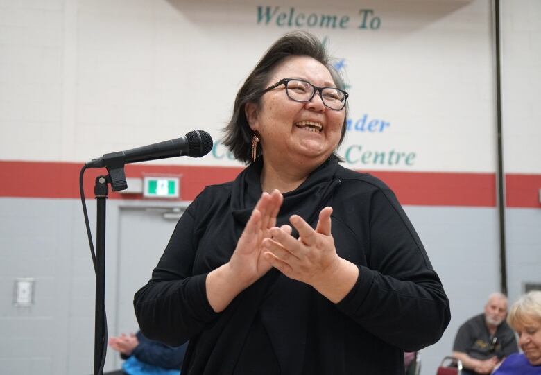 A woman smiles and claps her hands together in front of a microphone.