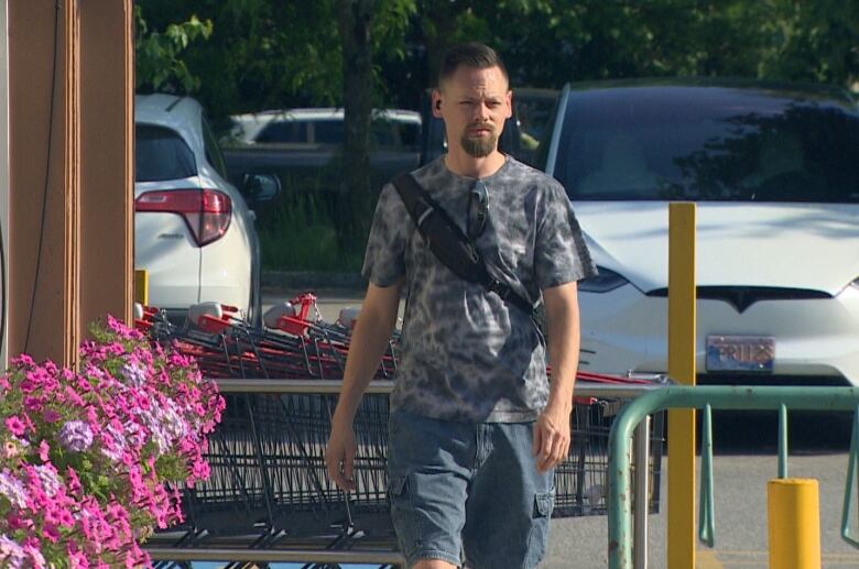 A white man with short brown hair and a brown beard walks in the sunlight in front of a parking lot. He is wearing a grey patterned t-shirt and blue cargo shorts.