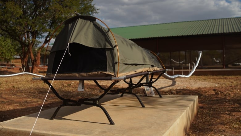 An army green single person tent is propped up on metal legs on top of a concrete pad with a long air conditioning like ducting tube connecting it to the enclosure where the scent is released. 