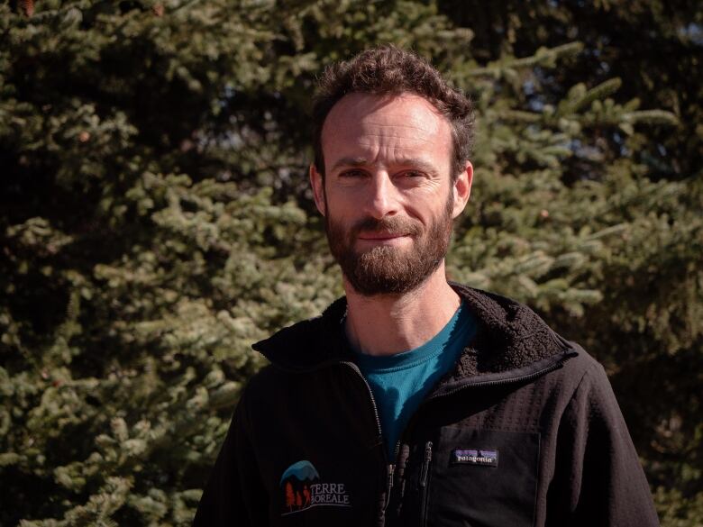 A bearded man poses in front of some trees.