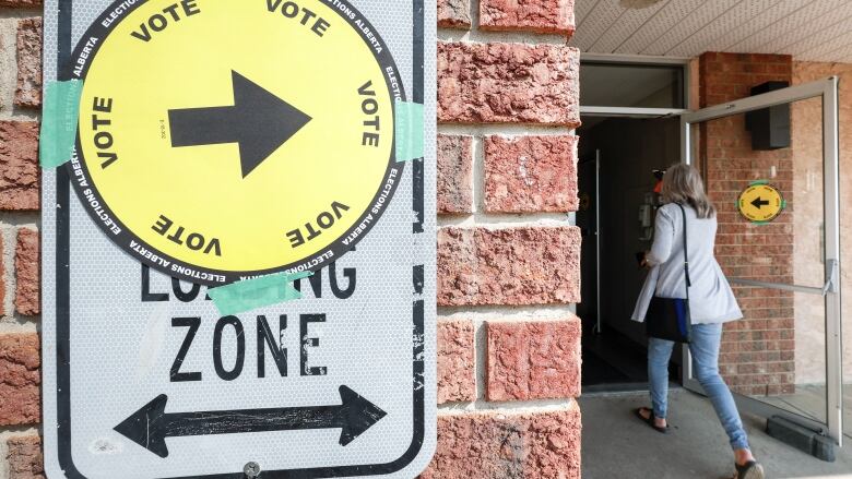 A woman with her back to camera walks through a glass doorway. At left, a yellow sign that covers a loading zone sign on the wall points to the voting station.