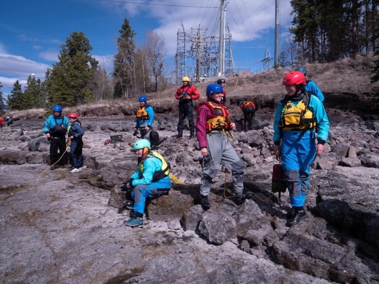 A group of people in helmets and lifejackets stand on a rocky shoreline,