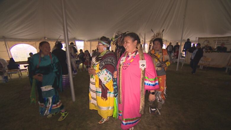 People dressed in traditional regalia smile with their arms locked together walking inside a tent at the powwow.