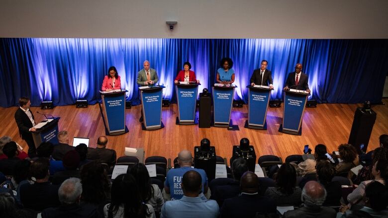 Six candidates stand at podiums on a stage.