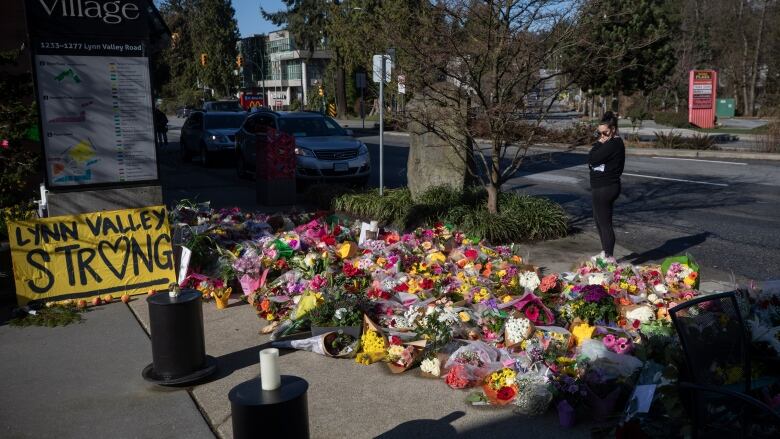 A woman dressed in black with dark hair and dark sunglasses stands in front of a pile of colourful bouquets laid in a pile on a concrete sidewalk on a sunny day. A yellow sign with 