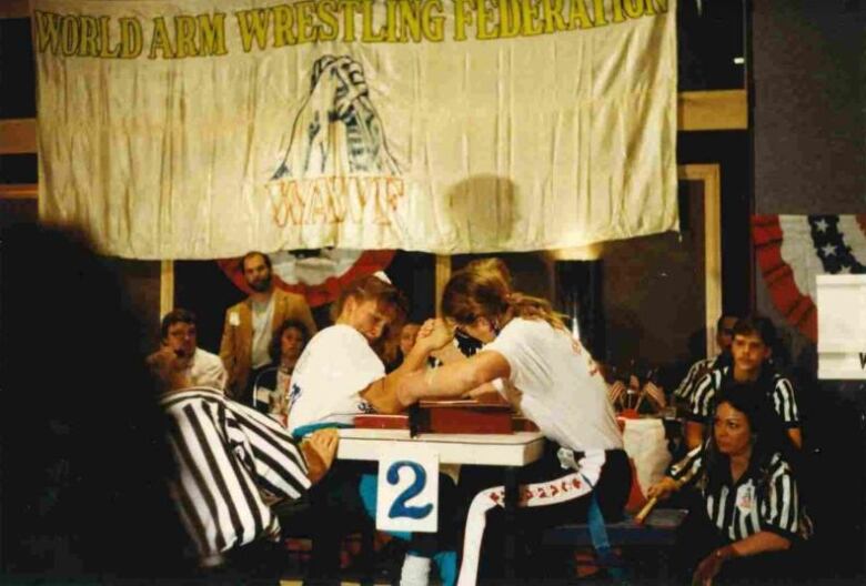 Two women arm wrestle surrounded by people in referee shirts, under a sign that reads,  'World Arm Wrestling Federation.'