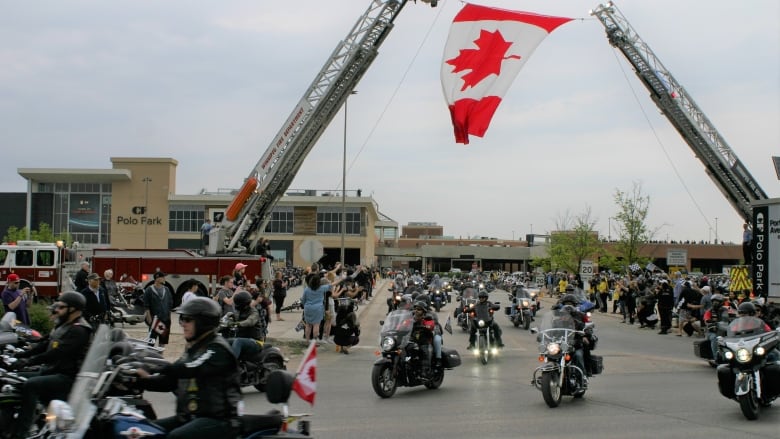 A large number of people riding motorcycles leave a parking lot. 