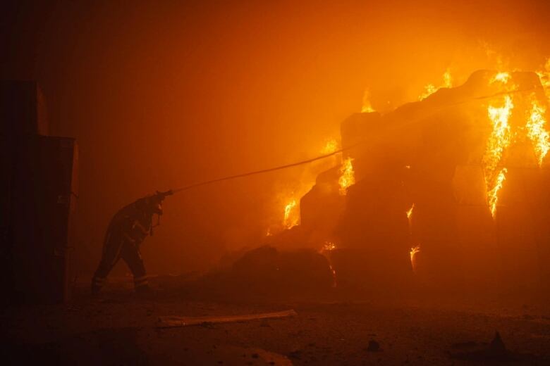 In a scene suffused with an orange haze, a firefighter sprays a fire taller than they are.