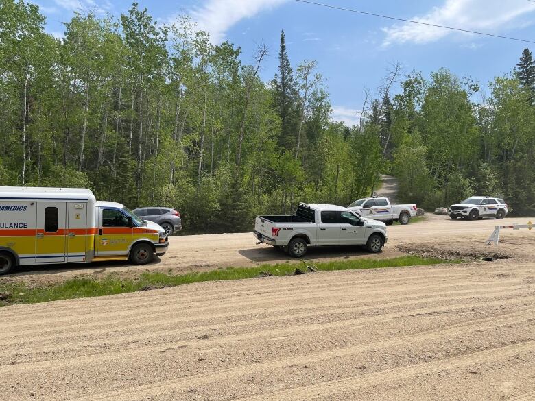 Two RCMP vehicles, an ambulance, a truck and a van are parked along the sides of a dirt road in a wooded area. 