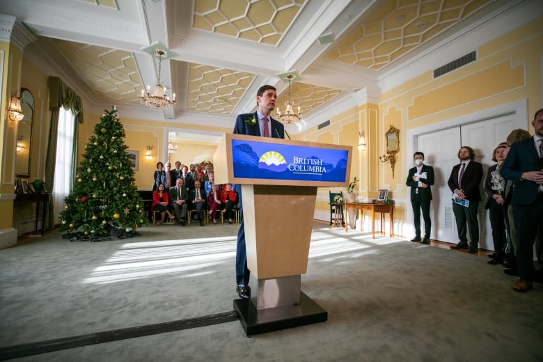 A white man stands in front of a Christmas tree and delivers a press conference at a podium.