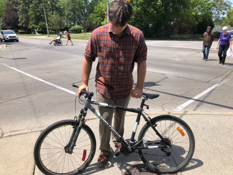 A man wearing a red plaid shirt and beige pants standing behind a bicycle