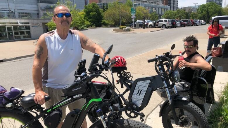 A man wearing a white tank top standing in front of a large bicycle and a man wearing a black tank top sitting on a bench beside another large bicycle