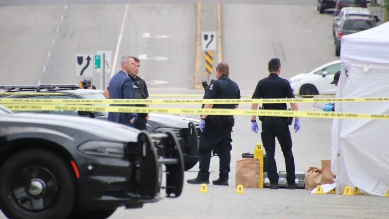 Police stand on a street, which has been cordoned off by police tape. A white tent covers a body and several vehicles and evidence markers sit on the street.