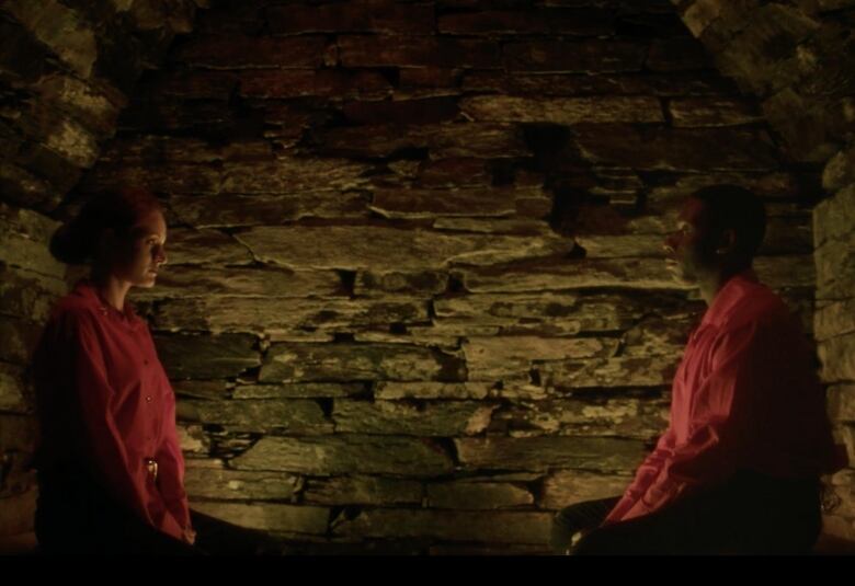 Two people sit facing each other in a dimly lit space with stone walls.