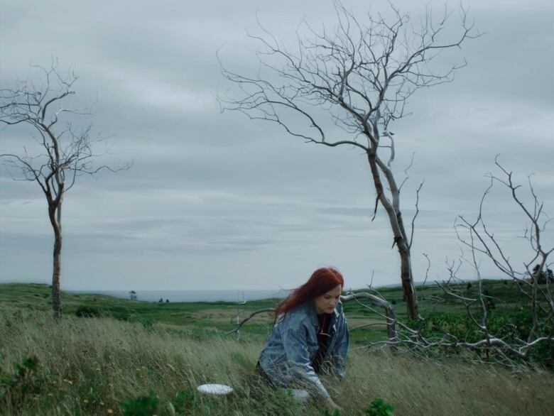 A long-haired woman in a denim jacket kneels in the grass, picking blueberries.