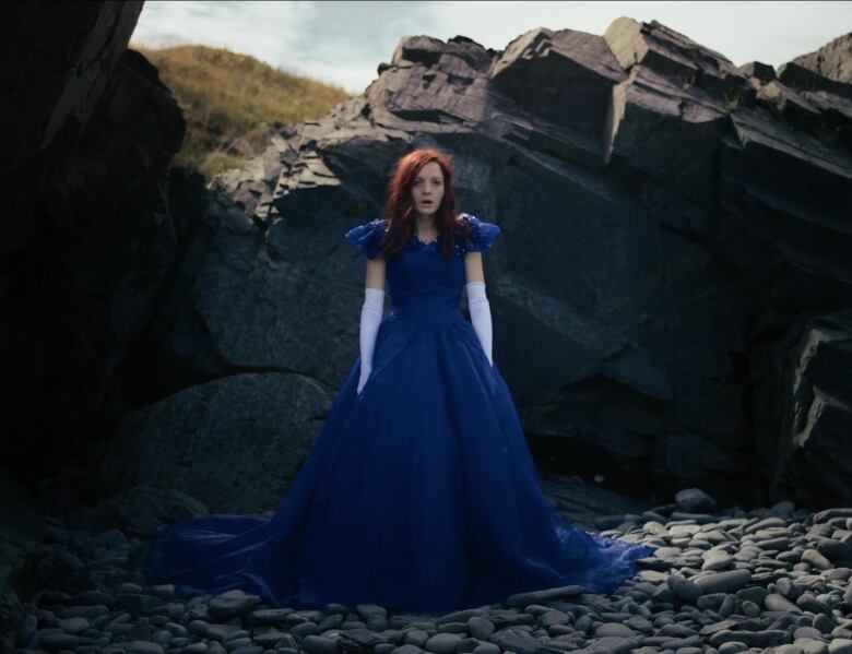 A woman in a flowing dress stands on a rocky beach on a sunny day.