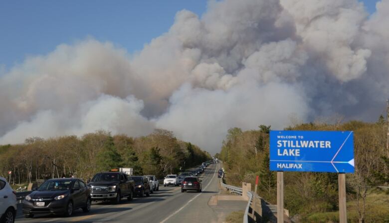 Cars on a road moving in the opposite direction of a wildfire whose heavy smoke can be seen in the distance in the sky.