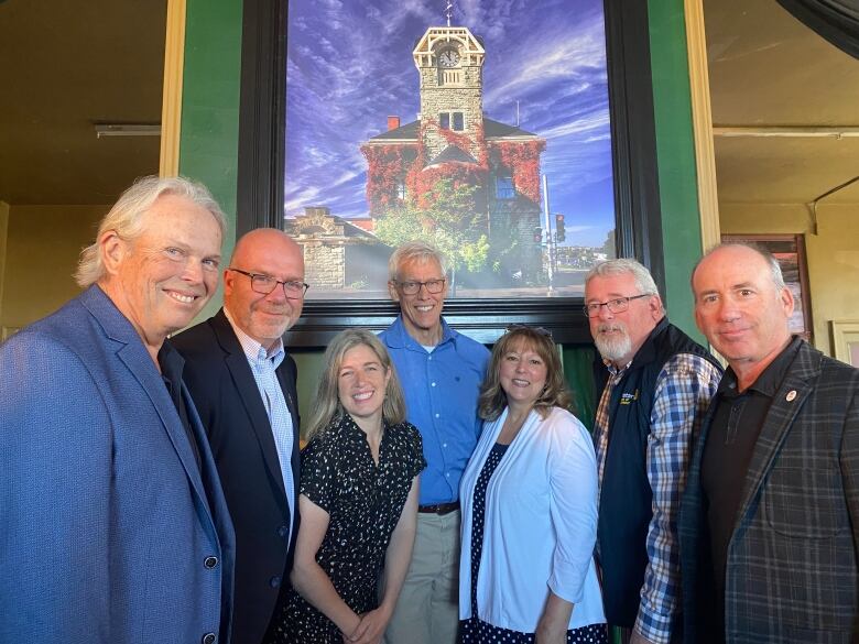 A group of seven people stand in a line smiling at the camera in front of a photo of an artistic building.