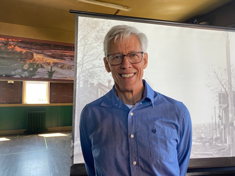 A man with short grey hair wearing dark-framed glasses smiles at the camera. He is wearing a denim-coloured button up dress shirt and standing in front of a projection of a historical black and white scene.