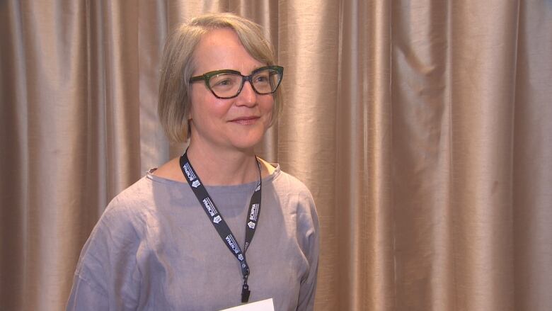 Woman stands in glasses, blue shirt and lanyard around her neck.