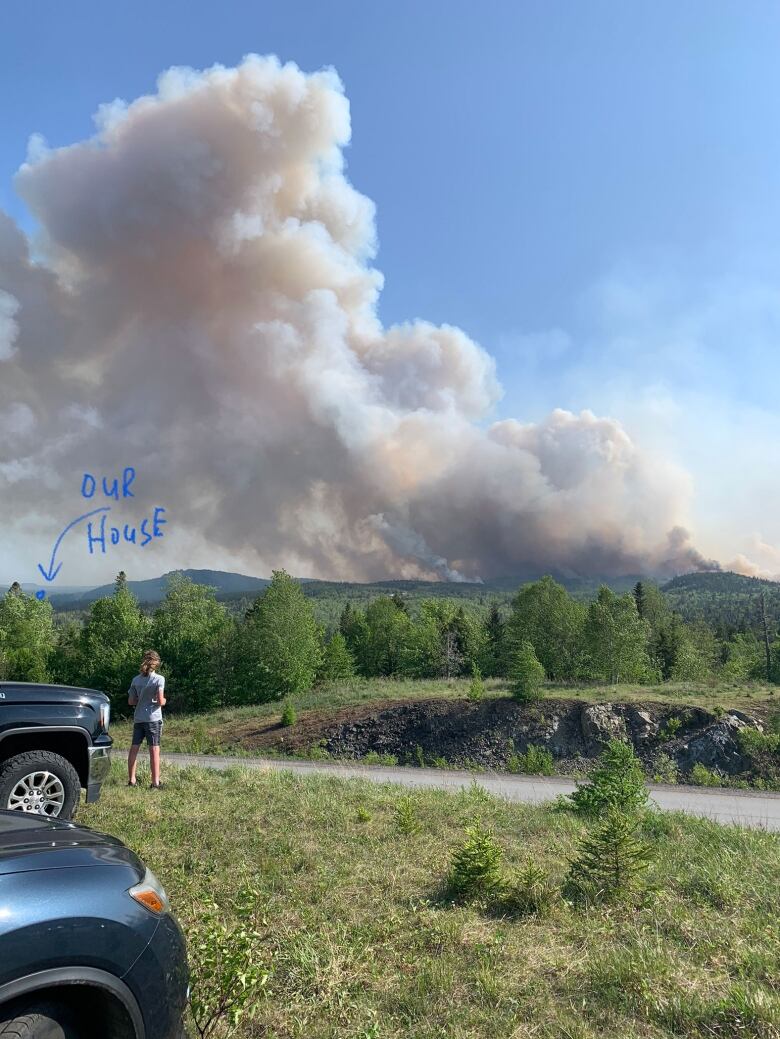 A person stands in front of a pickup truck, looking across the road toward some mountains, where thick white smoke from a forest fire is billowing high in the sky.