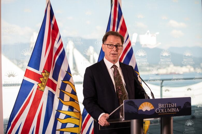 A white man talks at a podium, with two B.C. flags behind him.