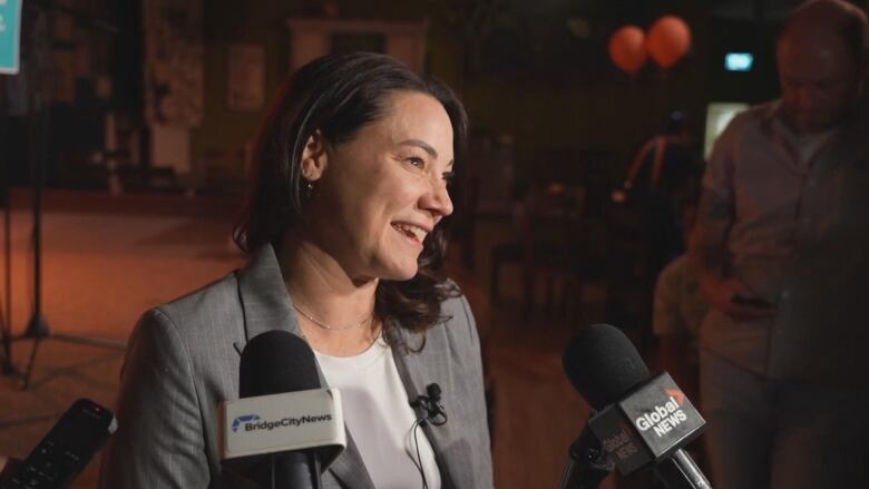 Shannon Phillips speaks into microphones and TV cameras in a dark room with orange balloons. 