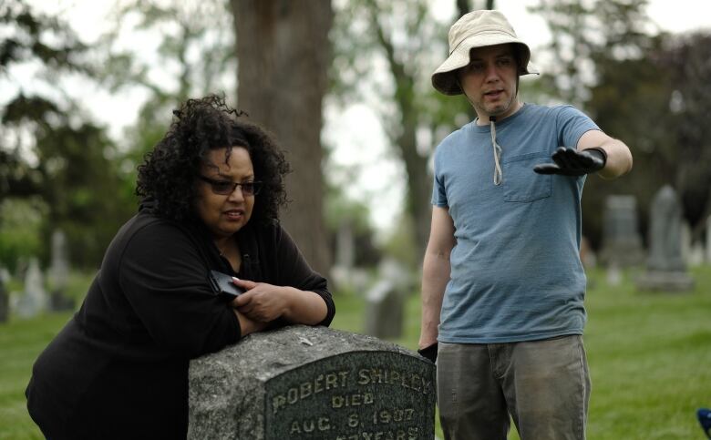 A Black woman leans on a gravestone while a white man stands beside her and gestures with his hand at something unseen in the graveyard.