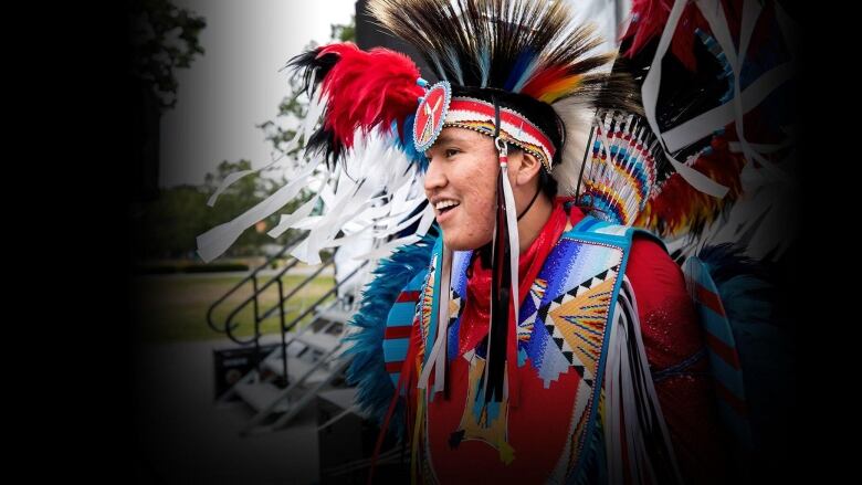 Indigenous man in a feathered headdress with plumes of red, white and black feathers and colourful shirt with varous Indigenous beading in gold, various colours of blue and white. 