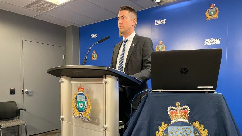 A man in a black suit stands at a podium with a coat of arms displayed prominently. He stands in front of a bright blue banner.